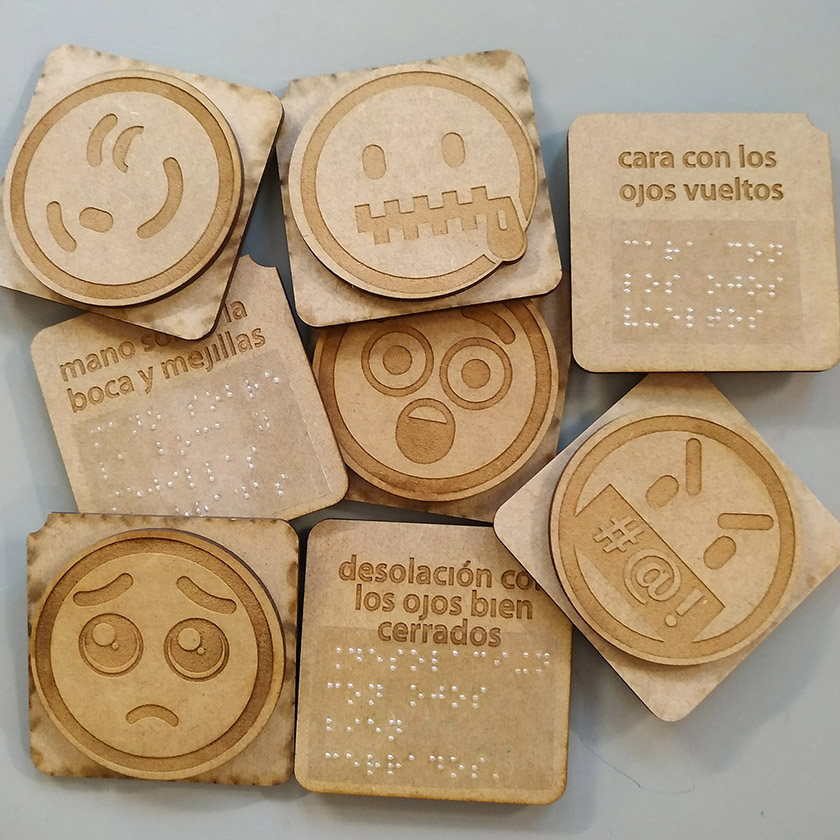 Varios emojis accesibles fabricados en madera, algunos vueltos del revés para que se vea la inscripción grabada en braille.